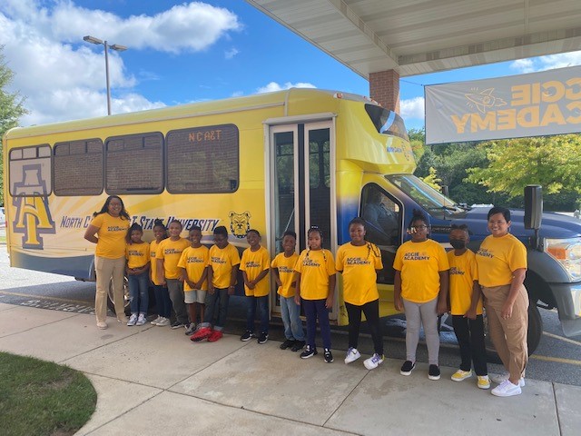 Elementary school students wearing yellow school shirts standing in front of a shuttle bus