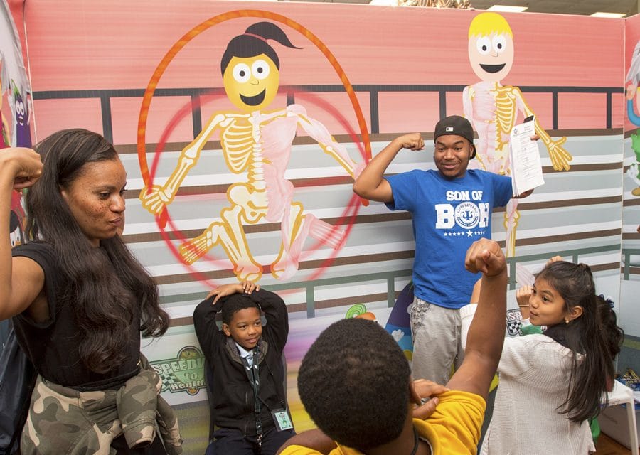 A group of children and adults flex their muscles in front of a colorful display showing cartoon skeletons inside animated characters. One man holds a paper while smiling, and others mimic strong poses in a playful, educational setting.