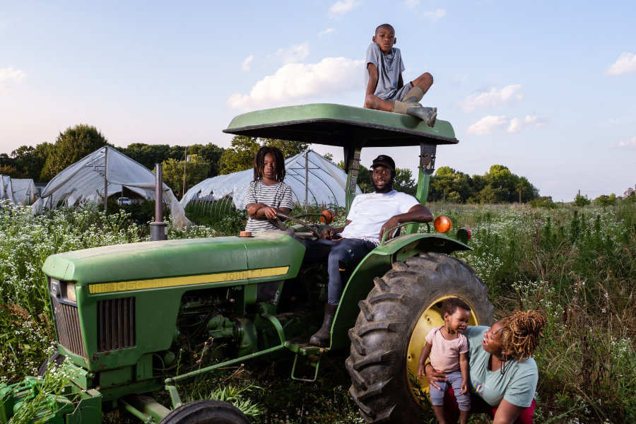 A family poses on and around a green John Deere tractor in a field of wildflowers. One child sits on top of the tractor’s canopy, another sits in the driver’s seat, and an adult man sits beside him. A woman crouches near the front tire, smiling at a toddler she is holding. Behind them are white plastic-covered hoop houses and trees under a blue sky with scattered clouds.