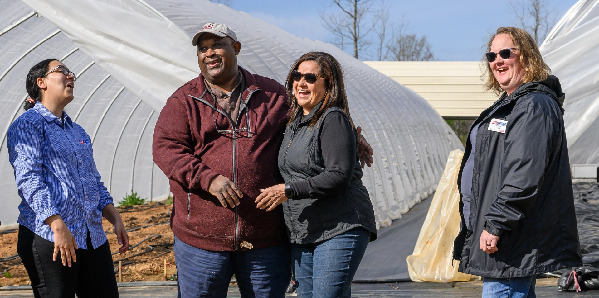Four adults stand laughing and smiling outdoors in front of plastic-covered greenhouses on a sunny day. One man and three women appear to be having a friendly conversation.